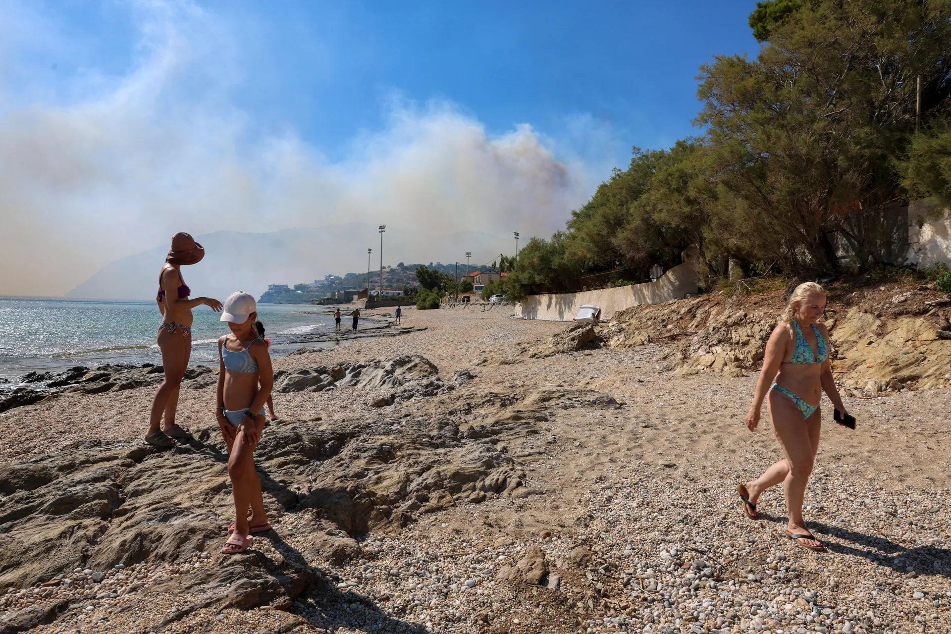 People walk on a beach as smoke rises from a wildfire burning next to the village of Plomari on the island of Lesbos, Greece, July 29, 2026. REUTERS/Elias Marcou