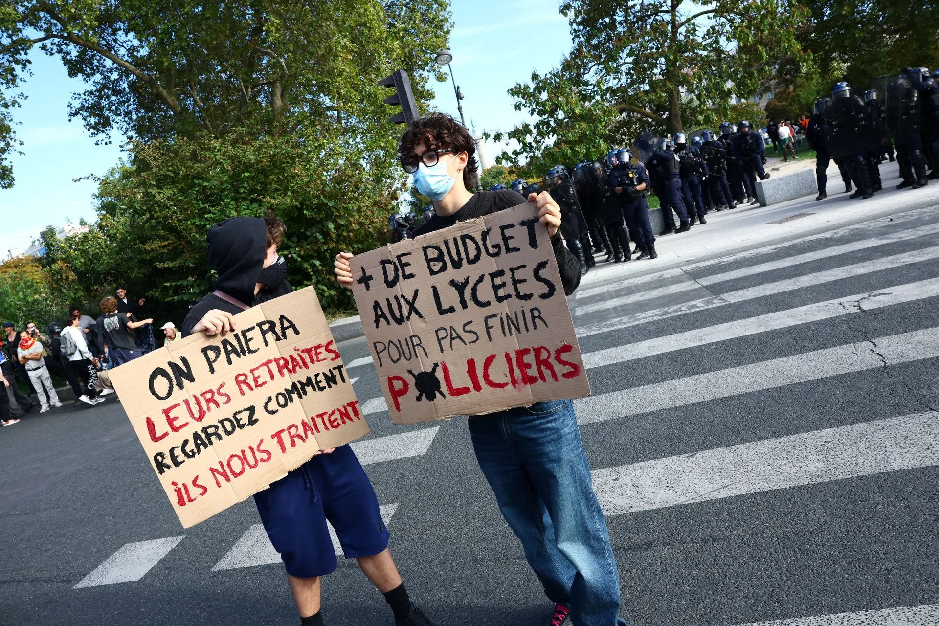 Protesters hold placards reading "We will pay for their pensions, look at how they treat us" and "More budget for high schools, so we don't end up as police officers" as French CRS riot police stand in position during a demonstration by high school and un
